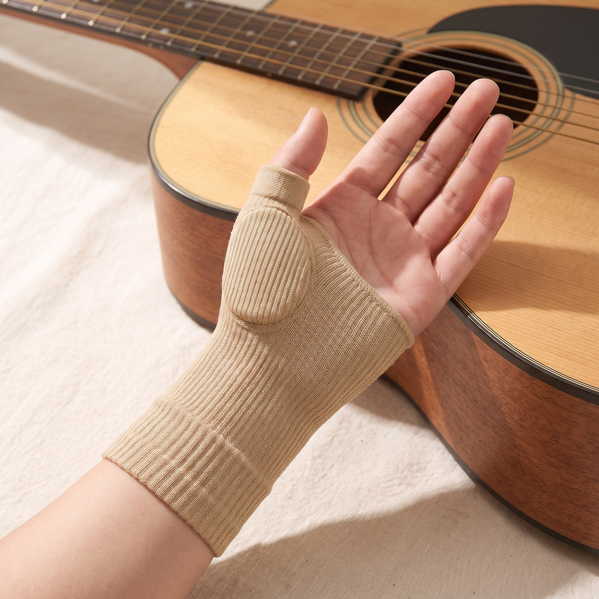 The beige knit thumb sleeve worn palm-up beside an acoustic guitar, showing the padding at the base of the thumb, the thumb tube, bare fingers and the ribbed cuff